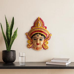 Puede incluir: Adorno de pared pintado a mano que representa el rostro de una deidad femenina. La obra de arte presenta un rostro color crema con labios rojos, un anillo en la nariz rojo y un tercer ojo. El tocado es amarillo y rojo.