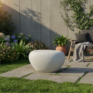 Könnte beinhalten: Ein weißer, kieselsteinförmiger Outdoor-Couchtisch aus Stein. Der Tisch steht auf einer Terrasse mit quadratischen Steinplatten und grünem Gras. Im Hintergrund befinden sich eine Betonmauer, Pflanzen und ein Holzstuhl.
