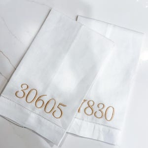Puede incluir: Dos toallas de mano de lino blanco con números dorados bordados. Una toalla muestra "30605" y la otra "7830". Las toallas están dobladas y colocadas sobre una superficie de mármol blanco.
