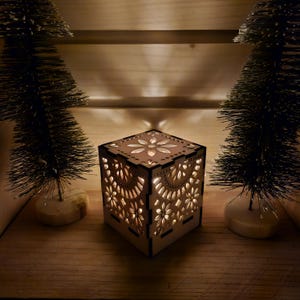 May include: A square, wooden candle holder with intricate floral cutouts. The candle holder is illuminated from within, casting a warm glow. Two small, artificial Christmas trees flank the candle holder, set against a wooden background.