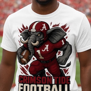May include: White t-shirt featuring a cartoon elephant in a Crimson Tide football uniform, holding a football. The elephant wears a helmet with the letter "A". The text "CRIMSON TIDE FOOTBALL" is at the bottom.