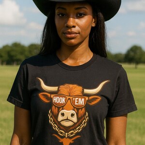 May include: Black t-shirt with a graphic of a longhorn wearing sunglasses and the text "HOOK 'EM". The person in the image is wearing a black cowboy hat and a brown belt.