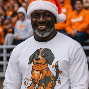 May include: A smiling person wearing a Santa hat and a white long-sleeve shirt. The shirt features a cartoon dog with an orange scarf and a letter "T" on it. The dog is decorated with Christmas lights. The background shows a crowd of people.
