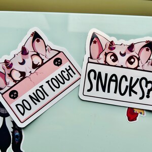 Puede incluir: Dos pegatinas con un gato de dibujos animados con orejas y cuernos rosas. Una pegatina tiene el texto "DO NOT TOUCH" sobre un fondo rosa. La otra pegatina tiene el texto "SNACKS?" sobre un fondo blanco.