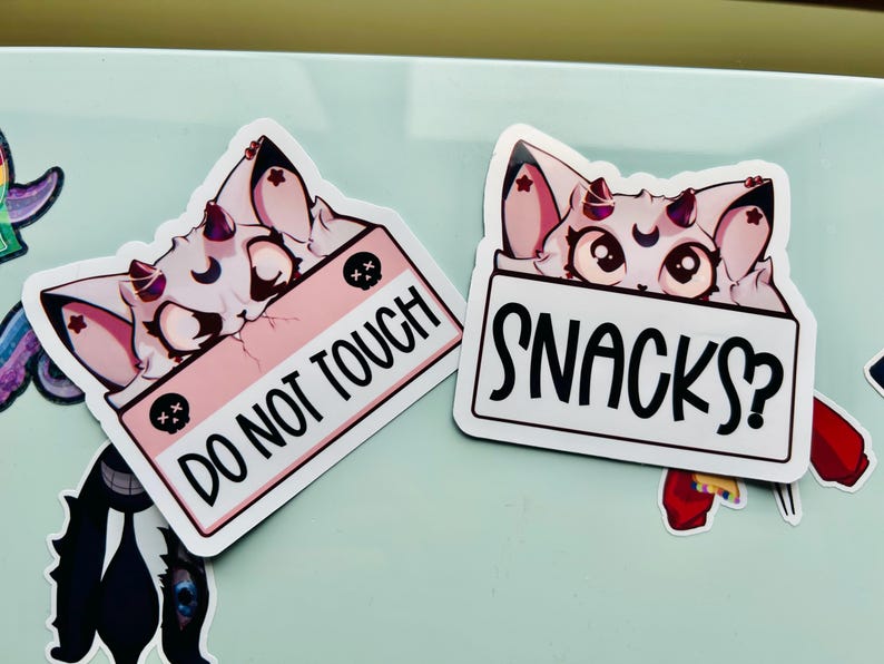 Puede incluir: Dos pegatinas con un gato de dibujos animados con orejas y cuernos rosas. Una pegatina tiene el texto "DO NOT TOUCH" sobre un fondo rosa. La otra pegatina tiene el texto "SNACKS?" sobre un fondo blanco.