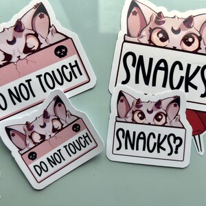 Puede incluir: Un conjunto de pegatinas con una criatura de dibujos animados con cuernos y una luna creciente. Las pegatinas muestran las frases "DO NOT TOUCH" y "SNACKS?" en texto negro sobre un fondo blanco.