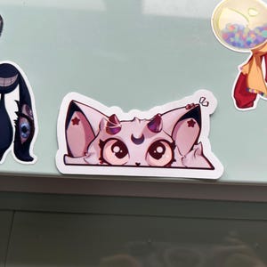Puede incluir: Una colecci&oacute;n de pegatinas coloridas sobre una superficie verde claro. Las pegatinas incluyen un gato de dibujos animados con pelaje rosa y blanco, una criatura negra con una cara sonriente, un dise&ntilde;o en espiral y una m&aacute;quina de chicles.