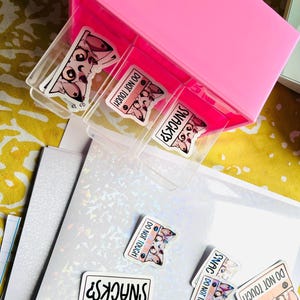 Puede incluir: Un organizador de pl&aacute;stico rosa con fundas de pl&aacute;stico transparente, que contiene pegatinas. Las pegatinas presentan un animal de dibujos animados con el texto "SNACKS?" y "DO NOT TOUCH". Algunas pegatinas est&aacute;n esparcidas sobre una superficie blanca iridiscente.