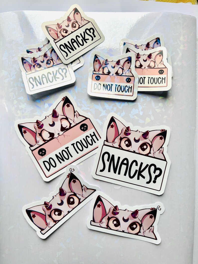 Puede incluir: Un conjunto de pegatinas con un gato de dibujos animados con cuernos y una luna creciente en la frente. Algunas pegatinas tienen el texto "SNACKS?" y otras "DO NOT TOUCH". Las pegatinas son blancas con detalles en rosa y negro.