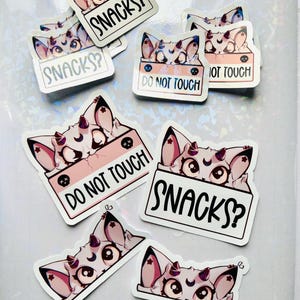 Puede incluir: Un conjunto de pegatinas con un gato de dibujos animados con cuernos y una luna creciente en la frente. Algunas pegatinas tienen el texto "SNACKS?" y otras "DO NOT TOUCH". Las pegatinas son blancas con detalles en rosa y negro.