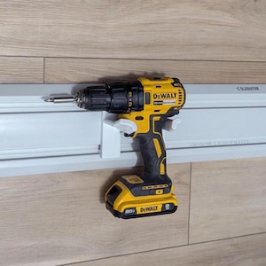May include: A yellow and black DeWalt cordless drill with a silver drill bit is mounted on a white holder. The drill is powered by a 20V battery. The drill is mounted on a white Gladiator wall storage system.