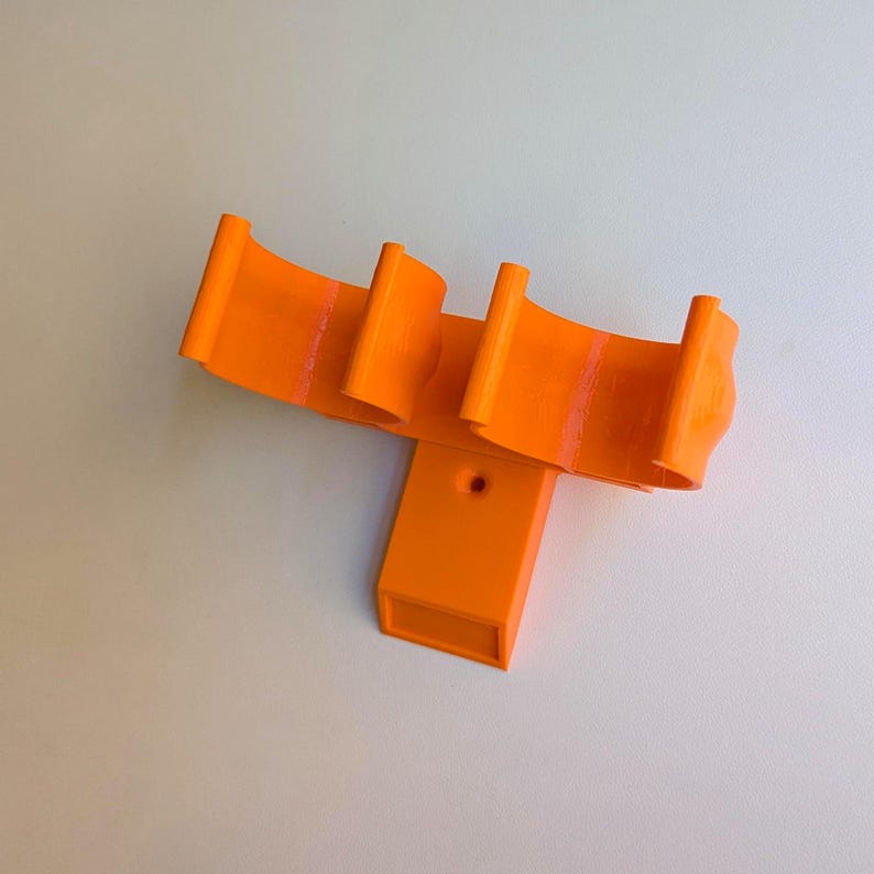 May include: Bright orange 3D-printed object with a unique design. It features a rectangular base with a hole and three curved, wing-like structures extending upwards. The object is made of solid orange plastic and appears to be a custom-made component.