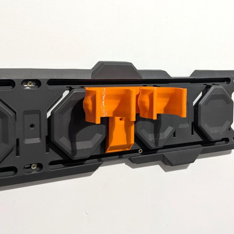 May include: A black wall-mounted storage system with orange brackets. The system has a rectangular shape with various geometric cutouts and screw placements. The orange brackets are designed to hold items, providing a functional and organized storage solution.