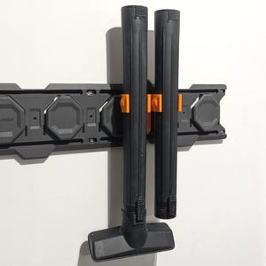 May include: A black vacuum cleaner attachment and two black tubes are held by orange clips on a gray wall-mounted storage system. The system is mounted on a white wall. The vacuum attachment has a rectangular head.