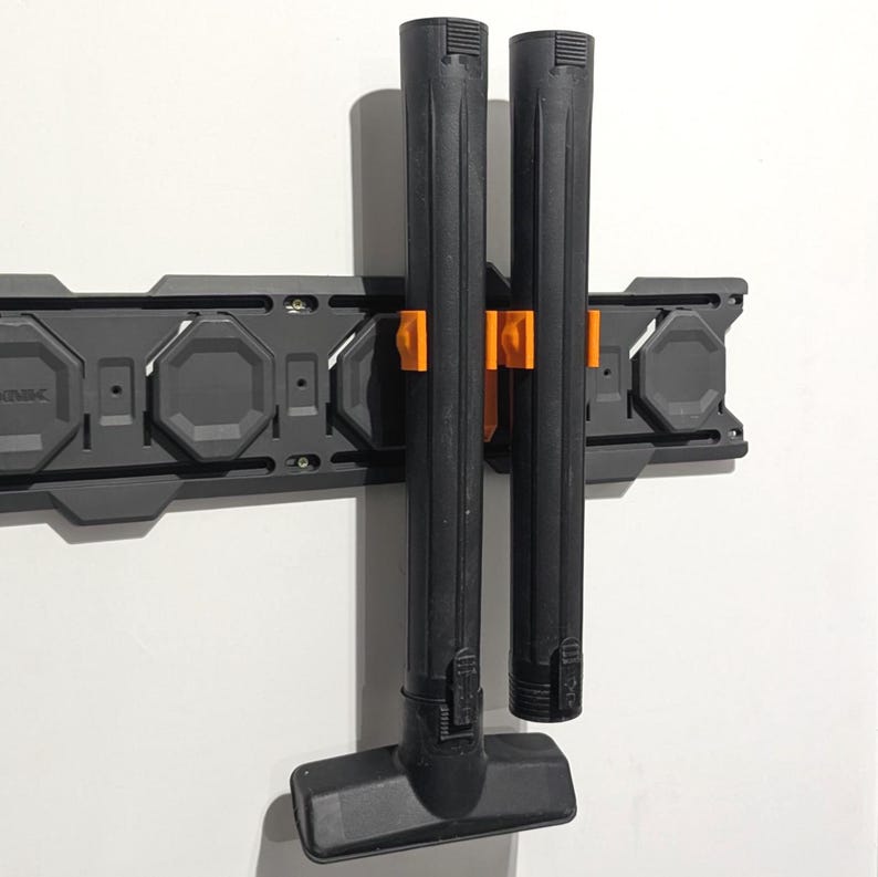 May include: A black vacuum cleaner attachment and two black tubes are held by orange clips on a gray wall-mounted storage system. The system is mounted on a white wall. The vacuum attachment has a rectangular head.