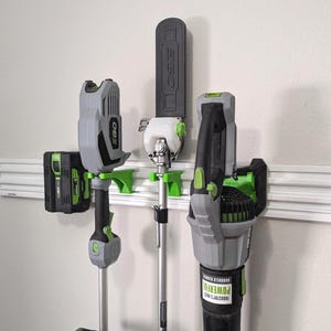 May include: A collection of gray and green cordless outdoor power tools, including a string trimmer, chainsaw, and leaf blower, mounted on a white wall. The tools are from the brand EGO and are designed for yard work.