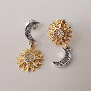 May include: A pair of celestial-themed earrings. One earring features a gold sun pendant with a central cluster of clear stones and a silver crescent moon. The other earring has a silver crescent moon and a gold sun pendant with clear stones.