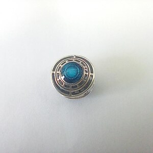 May include: A small, round, silver charm with a geometric pattern. The centre has a bright teal enamel inlay. The charm has a detailed, layered construction, creating a three-dimensional effect. The overall aesthetic is intricate and decorative.