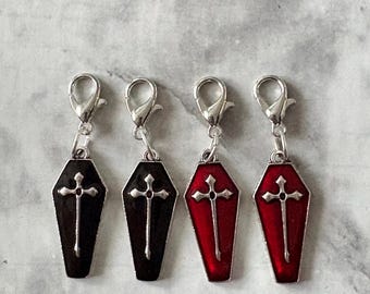 Op de afbeelding: Vier doodskistvormige bedels met zilverkleurige sluitingen. Twee zijn zwart met een zilveren kruis, en twee zijn rood met een zilveren kruis. De bedels hebben een zilveren rand.
