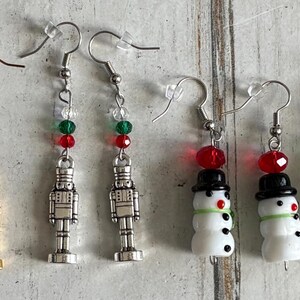 Puede incluir: Una colección de pendientes con temática navideña. Incluyen árboles de Navidad con detalles rojos y verdes, cascanueces dorados y plateados, y muñecos de nieve con sombreros negros. Los pendientes están en ganchos plateados.