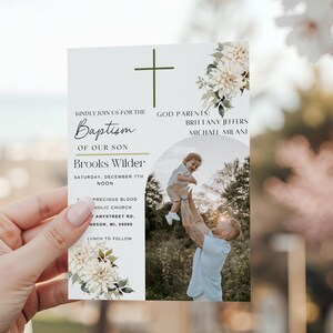 May include: A baptism invitation card held in a hand, featuring a photo of a person holding a child. The card has a cross and floral design, with text that reads "Baptism of our son Brooks Wilder."