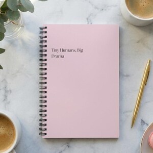 Puede incluir: Un cuaderno rosa con espiral con el texto "Tiny Humans, Big Drama" sobre una superficie de mármol, junto a un bolígrafo dorado, dos tazas de café, un plato de macarons rosas y un jarrón con vegetación.