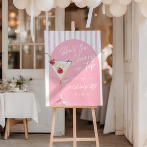 Puede incluir: Un letrero de bienvenida a rayas rosas y blancas sobre un caballete de madera. El letrero presenta una ilustración de cóctel con cerezas y el texto "She's the Cherry on Top" y "Welcome to Jackie's 40th Birthday". Globos blancos y cintas en el fondo.