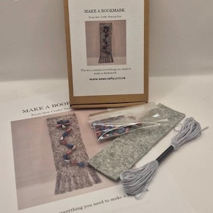 Puede incluir: Un kit de manualidades para hacer un marcapáginas, presentado en una caja de cartón con instrucciones impresas y una imagen de muestra. Incluye fieltro, cuentas, hilo y una hoja de instrucciones impresa. El texto en la caja dice "MAKE A BOOKMARK".