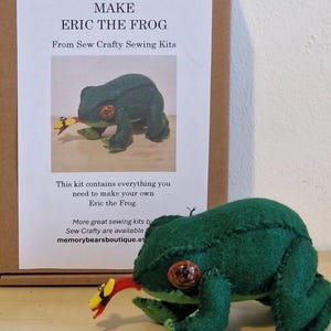 May include: A green felt frog with orange eyes and a red and yellow tongue. The frog is part of a sewing kit, with the text "MAKE ERIC THE FROG" on the packaging. The kit contains everything needed to make the frog.