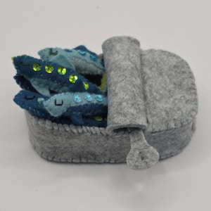 Puede incluir: Una lata de sardinas de fieltro con varios peces azules en su interior. Los peces tienen lentejuelas y son de color azul oscuro. La lata es gris y tiene una tapa de lengüeta.