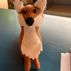 Puede incluir: Un juguete de zorro de fieltro hecho a mano. El zorro tiene un cuerpo marrón, pecho y cara blancos, y ojos y nariz negros. Las orejas del zorro son blancas con puntas negras. El juguete está de pie sobre una superficie azul.