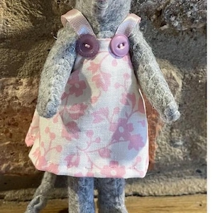 Puede incluir: Una muñeca de ratón de fieltro gris hecha a mano que lleva un vestido rosa con estampado floral y detalles de botones morados. El ratón tiene una nariz rosa, ojos negros y un lazo rosa. La muñeca tiene una larga cola gris y está de pie.