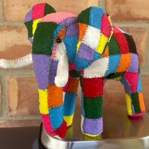 May include: A vibrant, handcrafted felt elephant toy. The elephant is made from fabric squares in pink, purple, green, blue, yellow, orange, and red. It features white tusks and a trunk, with hand-stitched seams. A patchwork design.