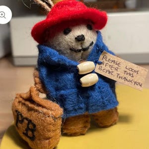 May include: A felt Paddington Bear ornament, wearing a red hat and blue coat with wooden toggles. The bear holds a brown satchel with "PB" on it and a tag that reads "Please look after this bear, thank you."