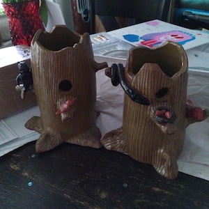 May include: Two brown ceramic tree stump planters with carved bark texture. Each has a hole and a small bird or animal figure attached. One has a black bear, the other has a black snake. The planters are on a table.