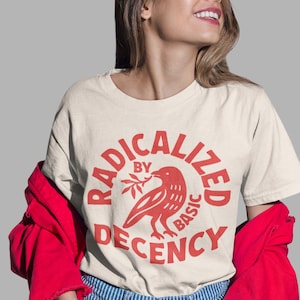 Op de afbeelding: Crèmekleurig T-shirt met een rode afbeelding van een vogel met een tak, omringd door de tekst "RADICALIZED BY BASIC DECENCY". De persoon draagt een rode jas en een blauwe gestreepte jeans.