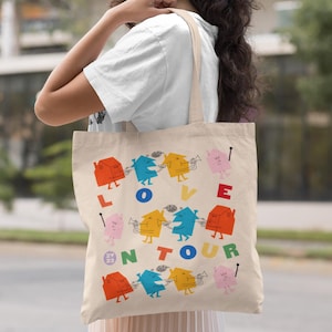 Pode incluir: Bolsa tote bege com um design gráfico colorido. O design apresenta casas de desenho animado tocando instrumentos musicais, com as palavras "LOVE ON TOUR 23" em uma fonte divertida. A bolsa tem uma forma retangular simples e uma alça de ombro.