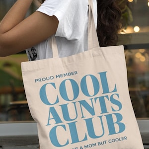 May include: A beige tote bag with the text "PROUD MEMBER COOL AUNTS CLUB LIKE A MOM BUT COOLER" in blue. The bag is carried over a person's shoulder, with a white t-shirt visible.