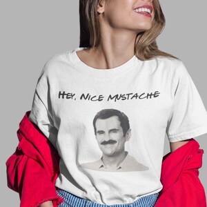 May include: White t-shirt with the text "Hey, Nice Mustache" above a black and white image of a man with a mustache. The person is wearing blue and white striped jeans and a red jacket.
