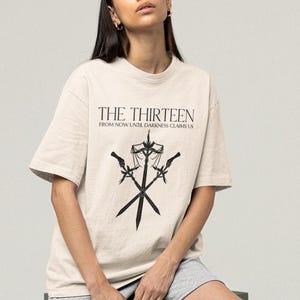 Pode incluir: Camiseta creme com o texto "THE THIRTEEN" e "FROM NOW UNTIL DARKNESS CLAIMS US" em preto. Abaixo, um gráfico preto de espadas cruzadas e uma balança.