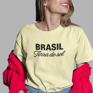 Puede incluir: Camiseta amarillo claro con las palabras "BRASIL Terra do sol" en negro. La persona lleva vaqueros a rayas azules y blancas y una chaqueta roja.