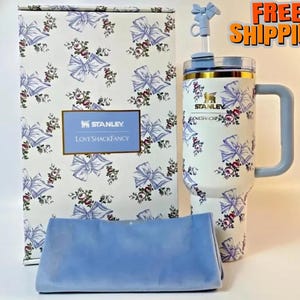 Puede incluir: Un vaso Stanley blanco con un estampado floral y de lazo, un asa azul y una pajita a juego. El vaso est&aacute; junto a una caja y un pa&ntilde;o azul, ambos con el mismo dise&ntilde;o. La caja tiene el texto "STANLEY LOVESHACKFANCY".
