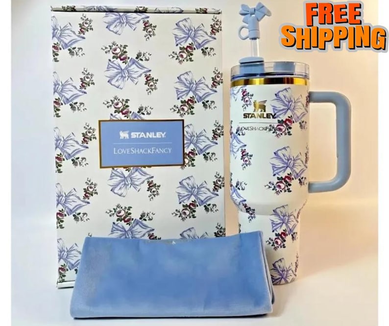 Puede incluir: Un vaso Stanley blanco con un estampado floral y de lazo, un asa azul y una pajita a juego. El vaso est&aacute; junto a una caja y un pa&ntilde;o azul, ambos con el mismo dise&ntilde;o. La caja tiene el texto "STANLEY LOVESHACKFANCY".