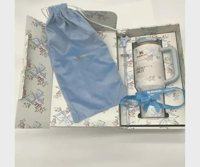 Puede incluir: Un juego de regalo con un vaso blanco y dorado con asa, una pajita azul y una cinta azul. El vaso est&aacute; decorado con motivos florales y las palabras "ENCHER TUMBLER". El juego incluye una bolsa con cord&oacute;n azul y est&aacute; empaquetado en una caja con estampado floral.