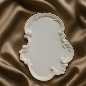 以下が含まれることがあります： 白い、装飾的なトレイ。縁は渦巻き模様で、滑らかな表面です。茶色のサテン生地の背景に配置されています。