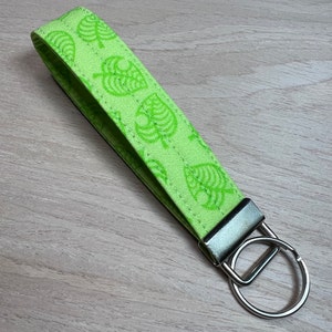May include: A green fabric keychain with a repeating pattern of white leaf designs. The keychain has a silver metal ring.