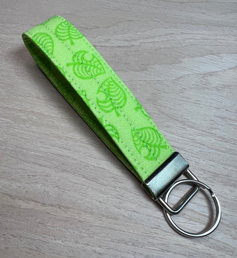 May include: A green fabric keychain with a repeating pattern of white leaf designs. The keychain has a silver metal ring.