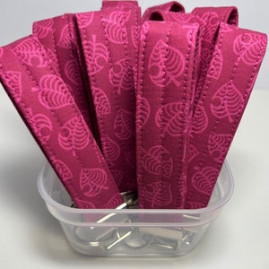 May include: A close-up of a container filled with pink fabric keychains featuring a white leaf pattern. The keychains are arranged in a stack and have silver key rings attached.