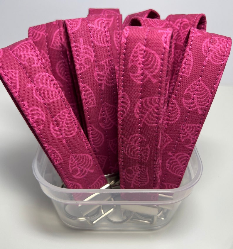 May include: A close-up of a container filled with pink fabric keychains featuring a white leaf pattern. The keychains are arranged in a stack and have silver key rings attached.