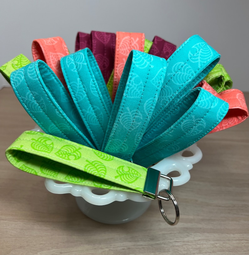 May include: A set of colorful fabric keychains with a leaf pattern. The keychains are in shades of green, blue, and pink. The keychains are arranged in a bowl.